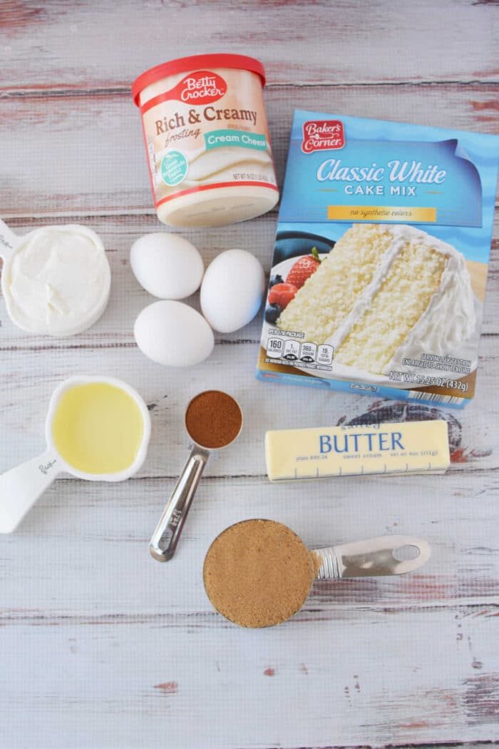 Cake mix, eggs, cream cheese frosting, butter, oil, sour cream, brown sugar, and cinnamon come together to create the solid cinnamon roll cake on a white table.