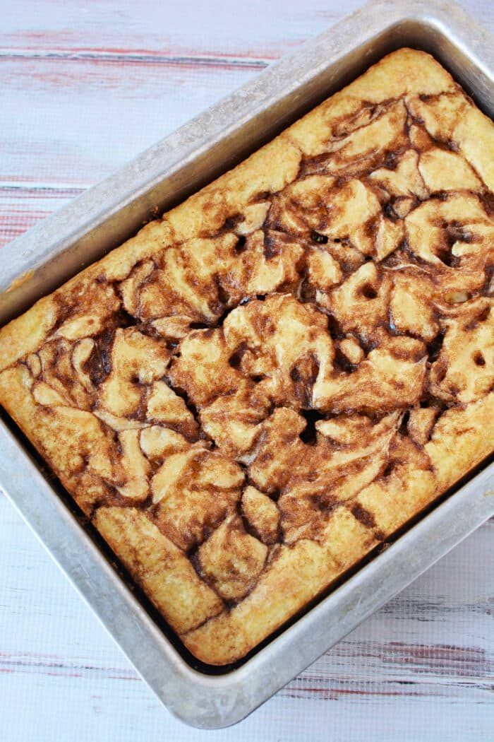 A Cinnamon Roll Cake with a beautiful cinnamon swirl pattern, baked in a rectangular metal pan using an easy cake mix, rests on a light wooden surface.