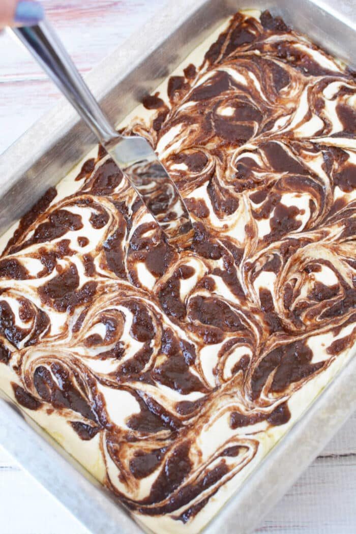 A hand swirls chocolate into batter with a knife in a metal baking pan, creating a marbled effect solid for a Cinnamon Roll Cake Recipe.