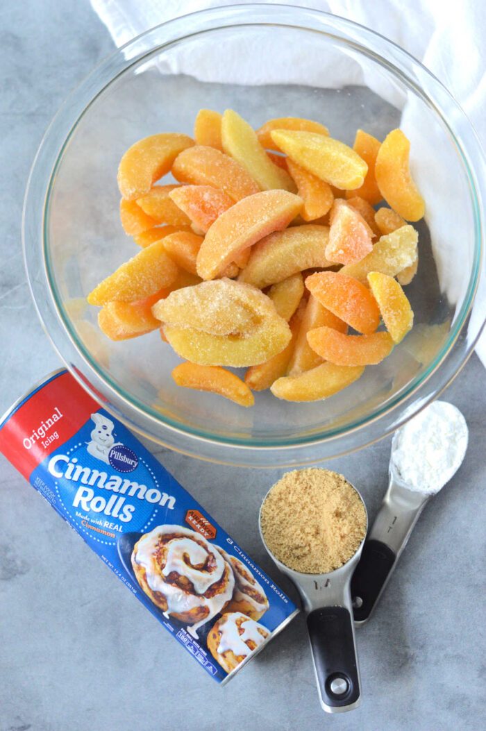 A bowl of frozen sliced peaches, cinnamon rolls can, brown sugar, and flour sit on a gray surface—solid for making an easy Cinnamon Roll Peach Cobbler.