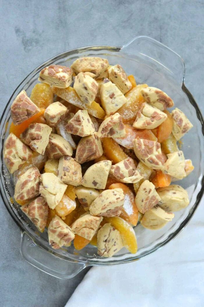 A glass pie dish with sliced peaches and chunks of marbled dough on a gray surface, this Cinnamon Roll Peach Cobbler promises a swirl of sweet, spiced flavor in every bite.