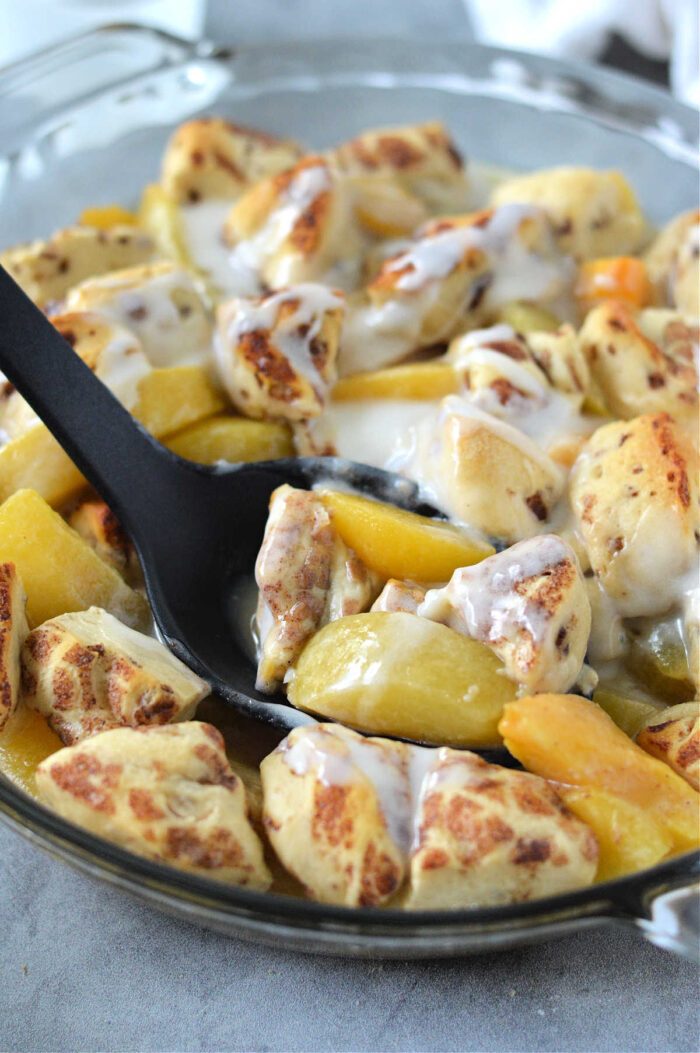 A baking dish of Cinnamon Roll Peach Cobbler with baked apples and cinnamon roll pieces, topped with icing and served with a black spoon.