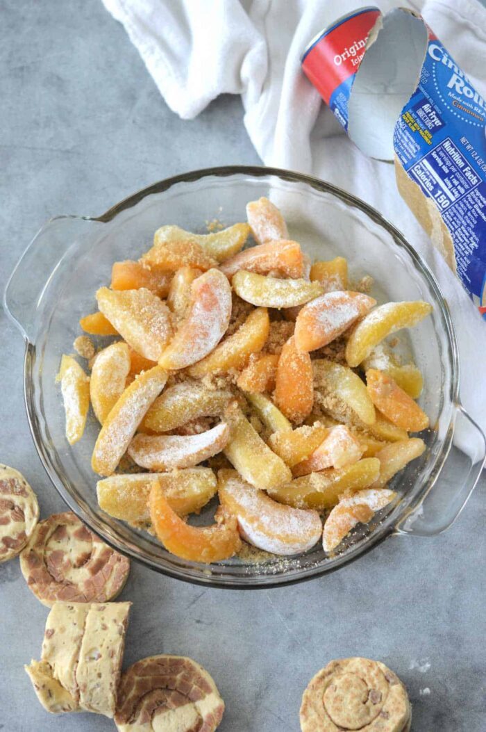 Sliced frozen peaches and apples with sugar in a pie dish, solid for making Cinnamon Roll Peach Cobbler, with biscuit dough and wrapper nearby on a gray surface.
