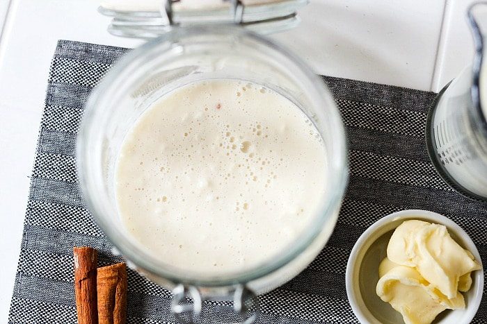 Top view of an open jar of frothy sourdough starter on a gray mat, evocative of a cinnamon roll recipe with no yeast, surrounded by fragrant cinnamon sticks and a bowl of rich butter nearby.