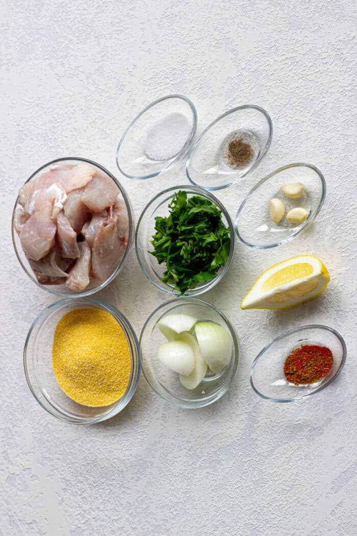 Ingredients for a dish: raw cod, herbs, lemon wedge, onion, spices, garlic, and cornmeal in small bowls. Prepare this mix with the same care you would use for crafting delicate Cod Fish Cakes.