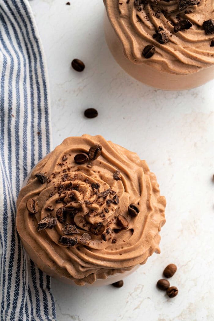 Two bowls of creamy chocolate coffee frosting, are topped with cocoa nibs and coffee beans on a white surface alongside a striped cloth.