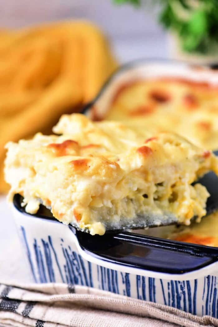 A slice of creamy baked mac and cheese cauliflower, reminiscent of a cheesy cauliflower bake, on a spatula above a blue casserole dish.
