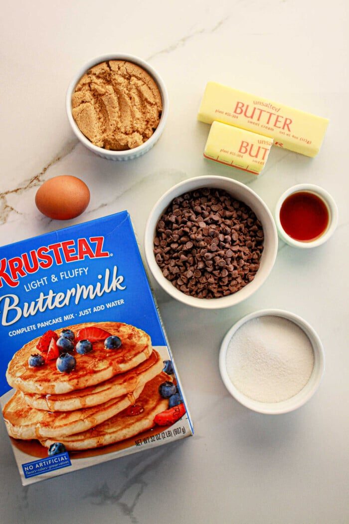 Ingredients for a baking recipe are laid out on a counter: a box of pancake mix solid for cookies, a bowl of brown sugar, chocolate chips, two sticks of butter, vanilla extract in a small cup, white sugar in another bowl, and one egg.
