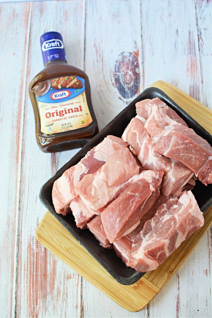 Raw boneless pork ribs rest on a black tray beside a bottle of Kraft Original Barbecue Sauce, solid for your next pork ribs recipe, all set on a wooden surface.