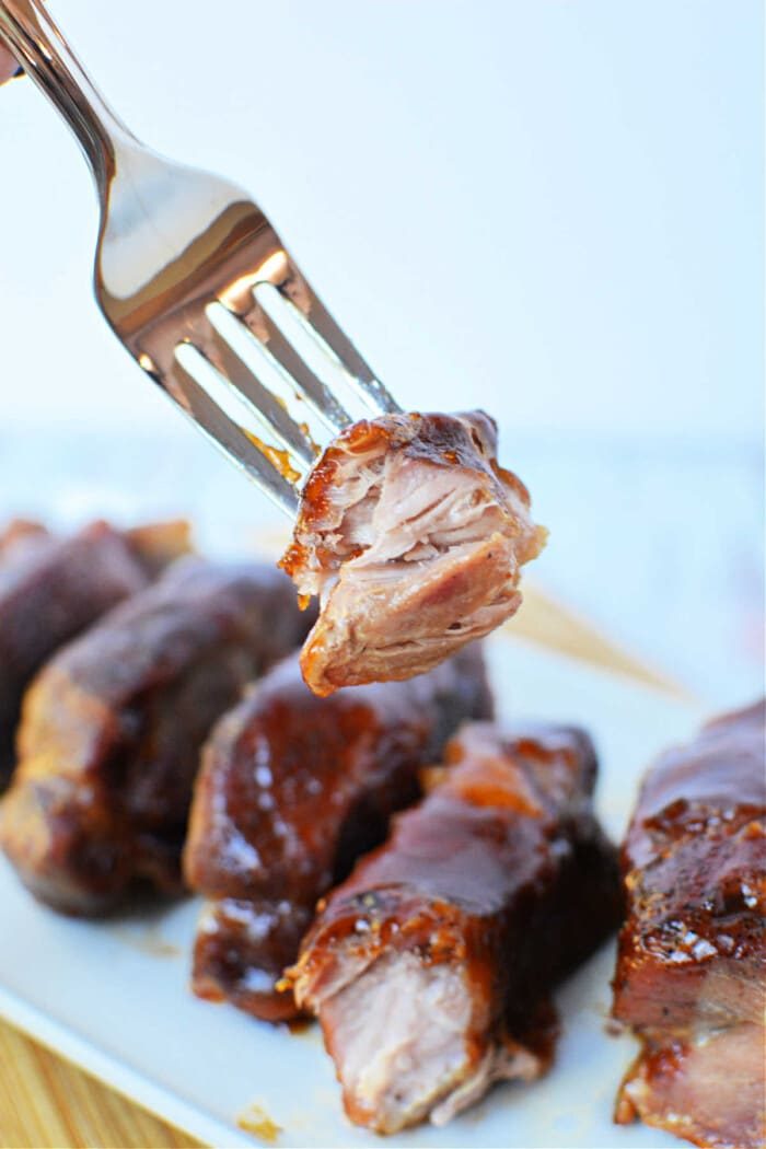 A fork holding a piece of glazed, tender boneless pork ribs above a plate of ribs, showcasing the solid result of oven cooking.