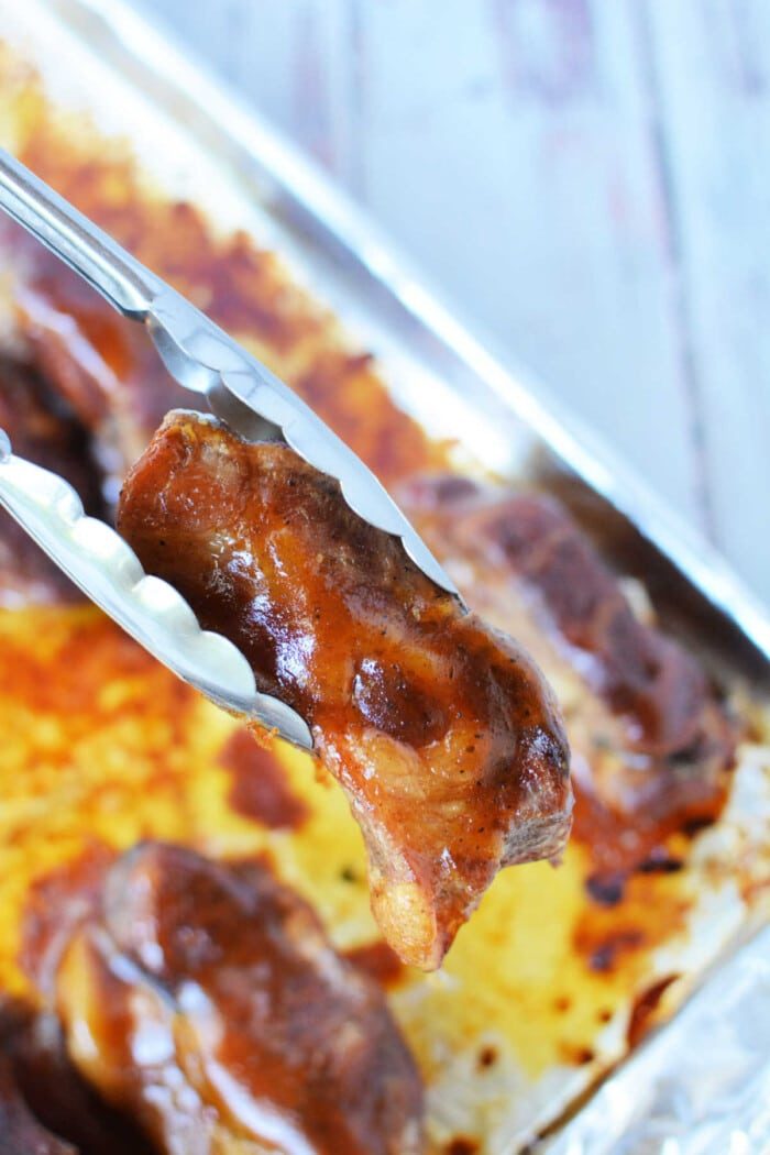 Tongs holding a piece of beautifully glazed boneless pork ribs above a baking tray, ready for the oven, with more succulent, glazed meat waiting beneath.