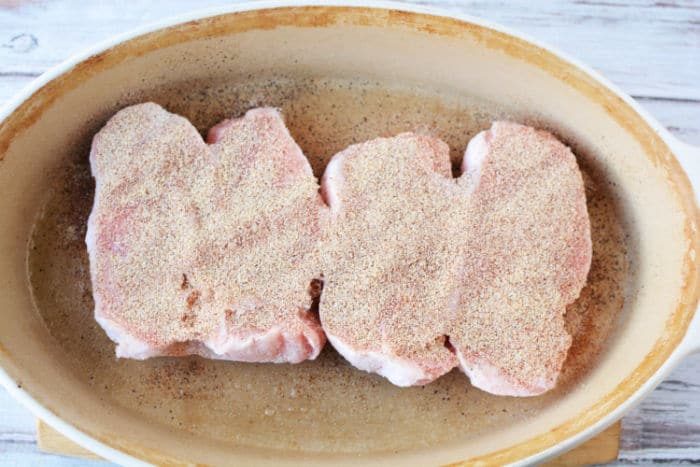 Three uncooked, seasoned frozen pork chops sit in an oval ceramic baking dish on a wooden surface, ready to be transformed into a good meal when baked in the oven.
