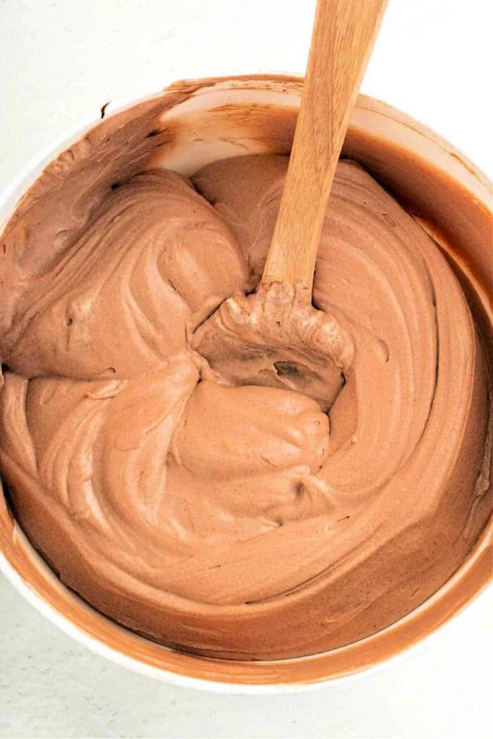 A bowl of creamy chocolate pudding frosting, enhanced with a dollop of Cool Whip, is being expertly stirred with a wooden spoon.