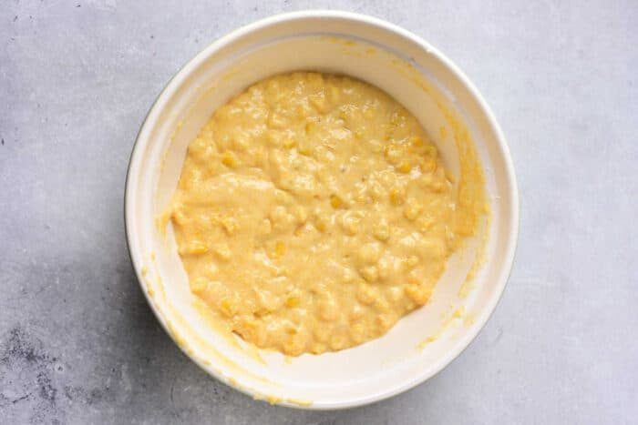 A white bowl filled with thick, yellowish batter containing visible corn kernels, placed on a light gray surface—solid for homemade Corn Pudding Without Jiffy Mix.