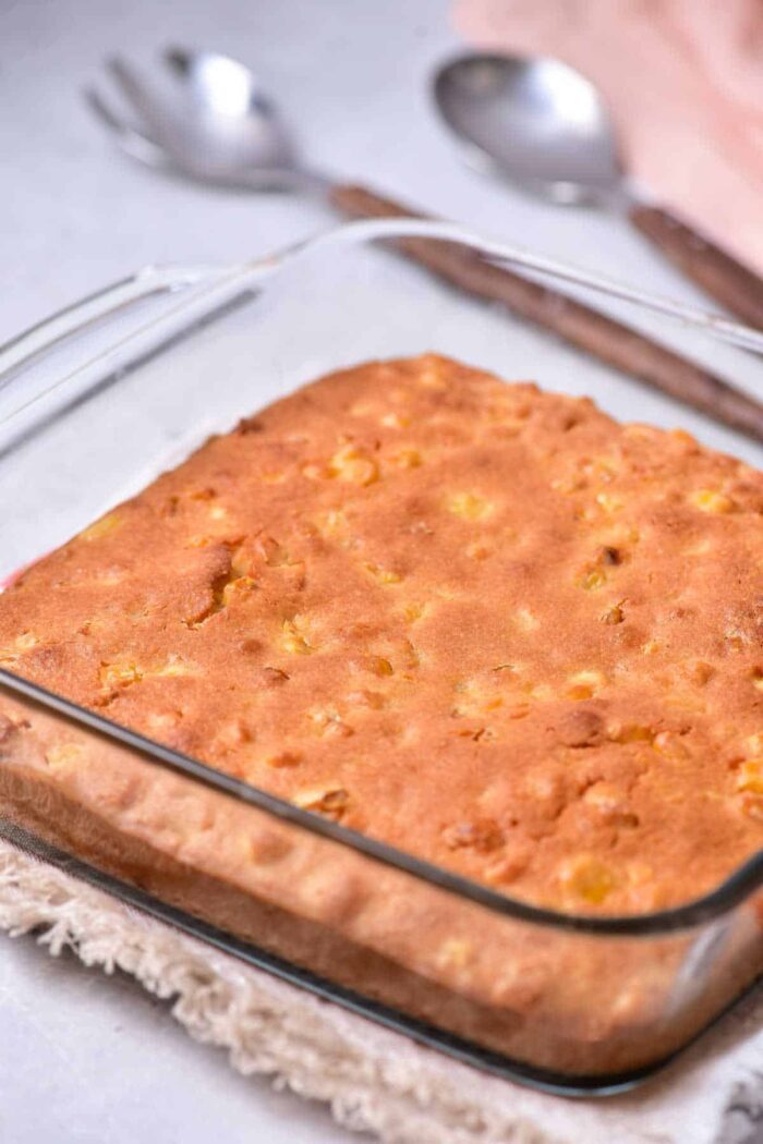 A square glass baking dish holds a freshly baked corn pudding with a golden-brown crust, reminiscent of homemade corn pudding without Jiffy mix. The dish is placed on a tan woven trivet, and two silver spoons are in the background on a light-colored surface.