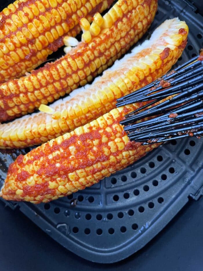 Corn on the cob ribs, brushed with vibrant red seasoning, transforms into a good corn ribs recipe as it cooks in a sleek black air fryer basket.