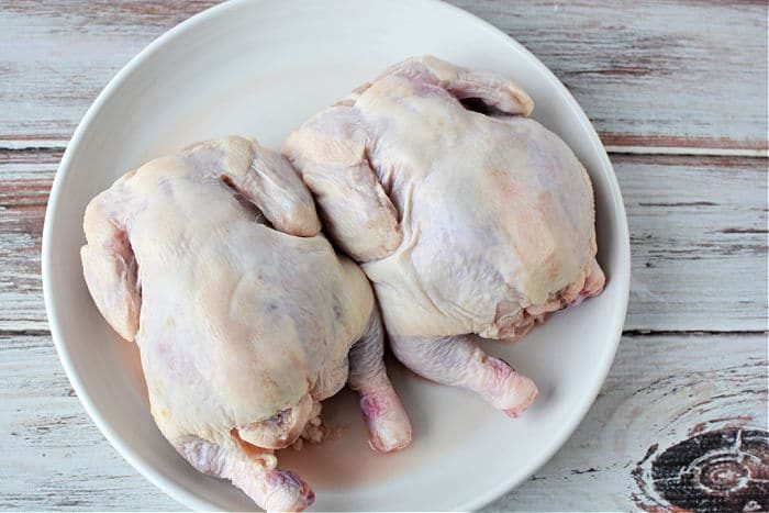Two raw whole game hens rest side by side on a white plate atop a rustic wooden surface, evoking questions like how long to cook Cornish hen in an air fryer for the solid meal.