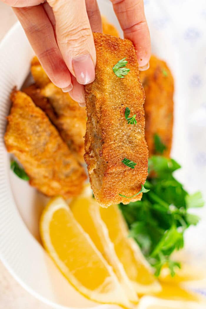 A hand holds a crispy fried piece of fish, perfectly coated in cornmeal for that extra crunch, over a plate adorned with lemon wedges and fresh greens.