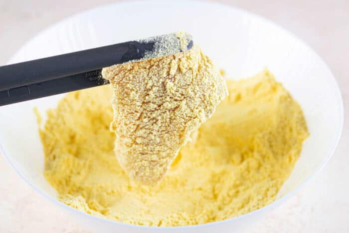 Black tongs hold a piece of food coated in seasoned yellow cornmeal for a fish fry, above a bowl filled with the same cornmeal mixture.
