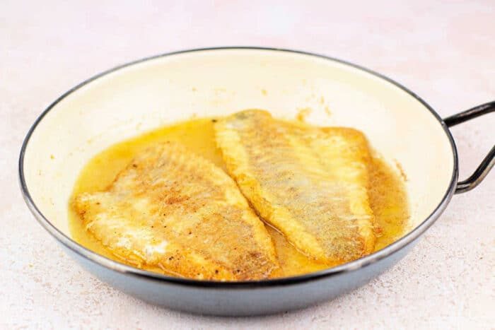 Two cooked fish fillets in a frying pan, coated in light brown sauce and cornmeal mixture. The fillets are golden brown, partly submerged in sauce.