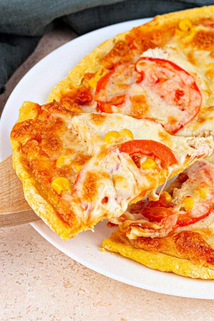 Close-up of a cheesy pizza slice with tomatoes on a cornmeal pizza crust being lifted from the whole pie on a white plate.