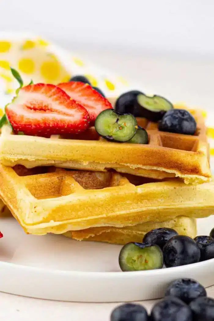 A plate with two cornmeal waffles topped with sliced strawberries and blueberries. A yellow and white polka dot cloth is in the background.