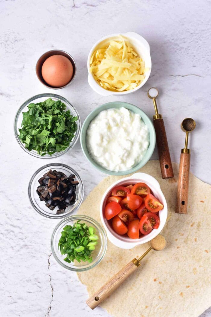 A flat lay image showcases various ingredients for a meal, solid for making Cottage Cheese Quiche. These include grated cheese, an egg, chopped spinach, ricotta or cottage cheese, cherry tomatoes, chopped mushrooms, green onions, and a tortilla. Two wooden-handled scoops are placed nearby.