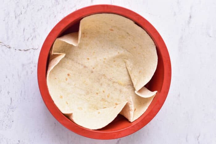 A single tortilla is arranged inside a red, round baking dish on a light-colored surface. The edges are slightly folded and overlapping, forming a semi-crumpled appearance, solid for a good cottage cheese quiche. The background is textured with subtle cracks and lines.