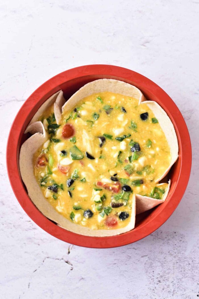 A red bowl filled with a yellow cottage cheese quiche mixture, brimming with chopped vegetables like olives, tomatoes, green peppers, and corn. The bowl is lined with tortilla pieces. The background is a white, textured surface.