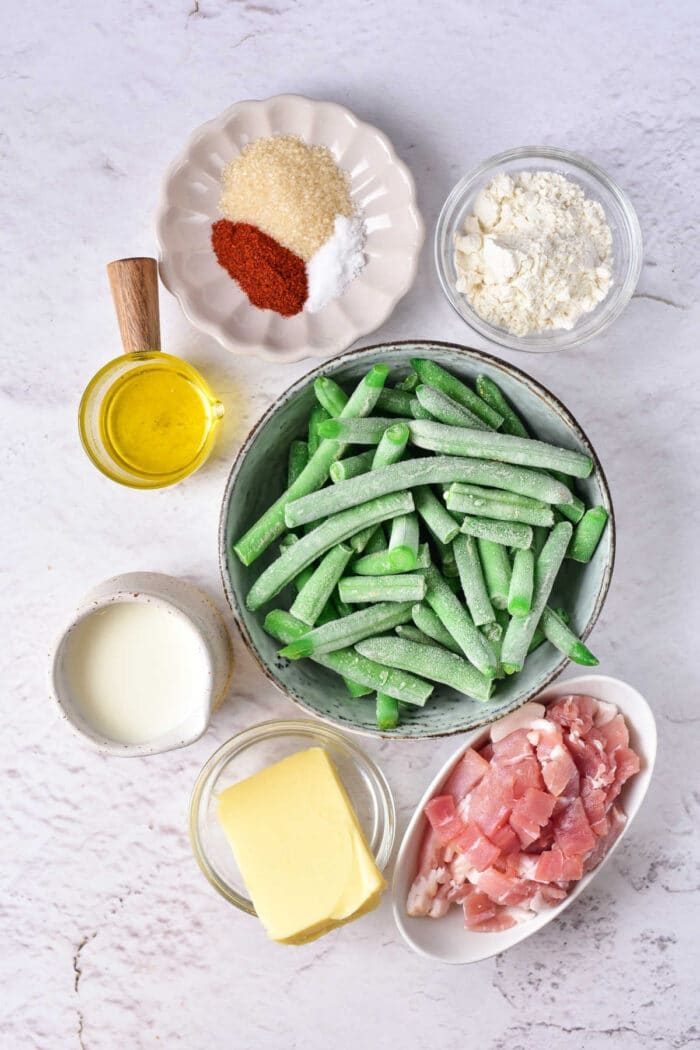 Ingredients on a table: crack green beans, bacon, butter, milk, flour, spices, oil, and brown sugar neatly displayed in small bowls.