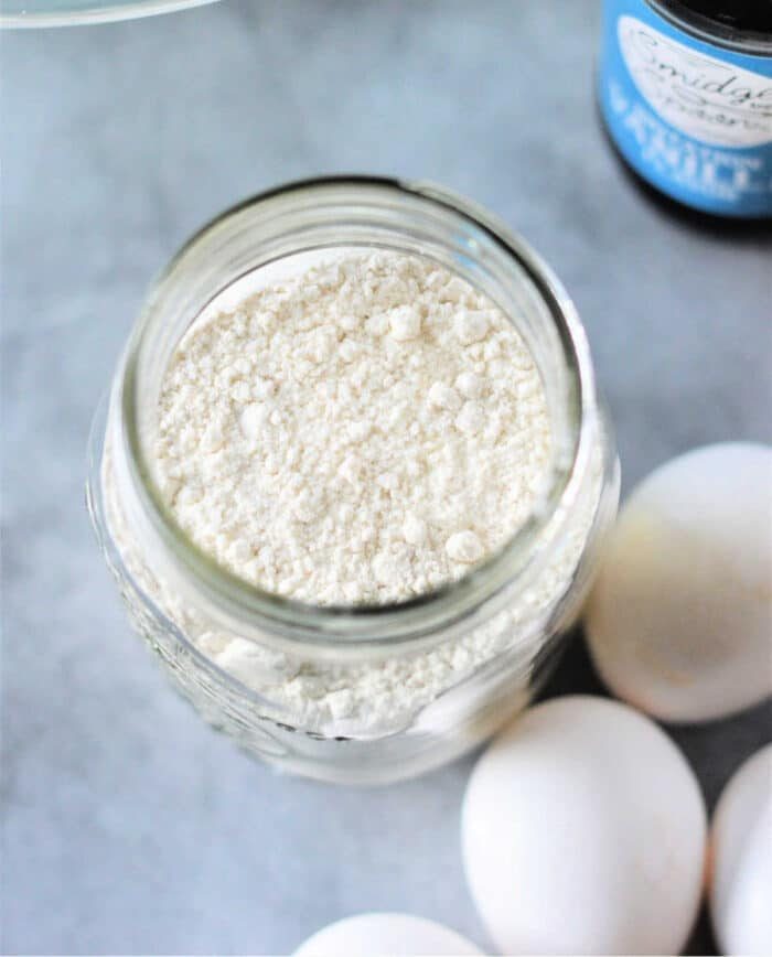 A jar filled with pancake mix sits on a surface, surrounded by eggs, as if preparing for something good, like Cracker Barrel Pancake Mix. A bottle is partially visible in the background.