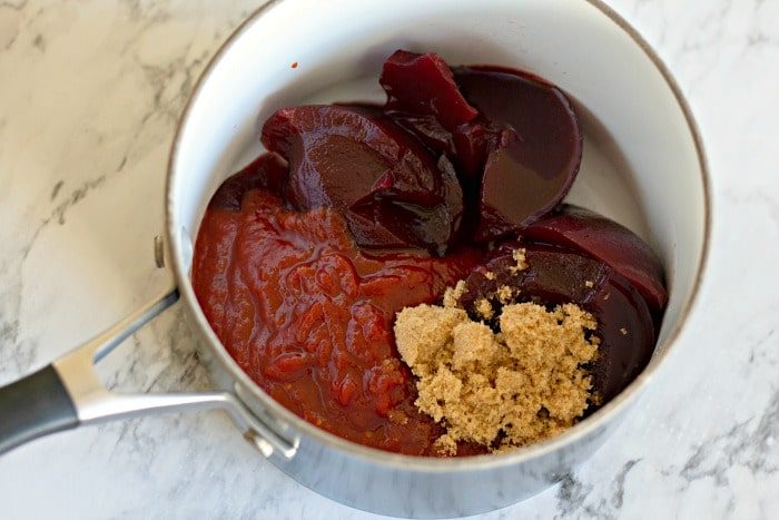 A saucepan with sliced cranberry sauce, ketchup, and brown sugar sits on a marble countertop, ready to enhance your cranberry meatballs with its tangy sweetness.