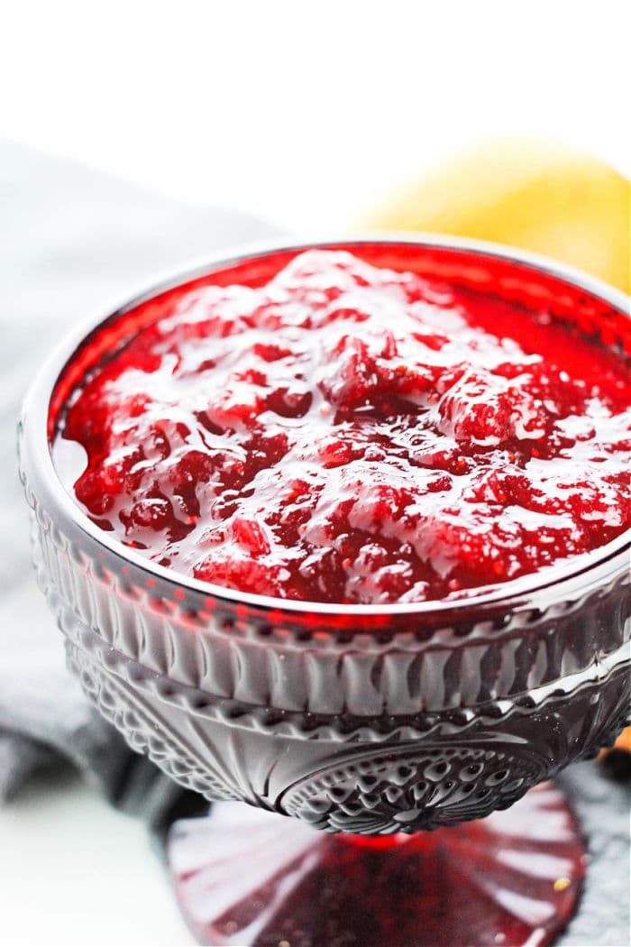 A glass bowl of vibrant cranberry sauce recipe with hints of orange juice, shines with textured details against a pristine white background.