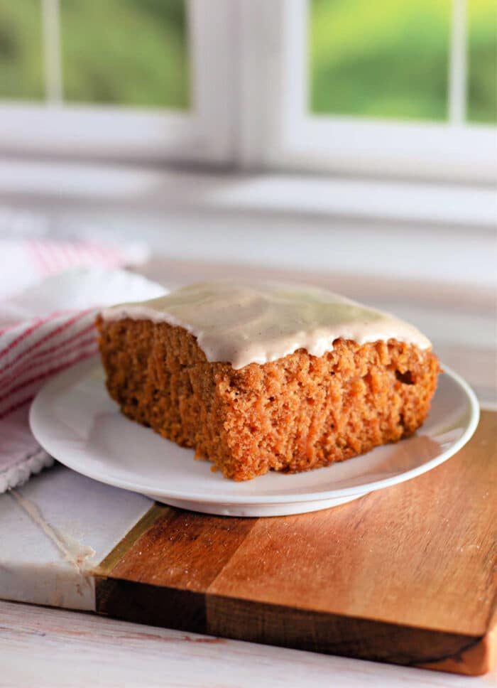 A slice of frosted Crazy Cake carrot cake sits on a white plate. The cake has a thick, soft texture and is topped with a light layer of white icing. The plate rests on a wooden board, with a white and red striped cloth partially visible in the background.
