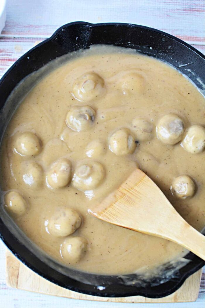 A skillet filled with succulent mushrooms smothered in a creamy brown sauce enriched with the flavors of cream of mushroom gravy, skillfully stirred with a wooden spatula.