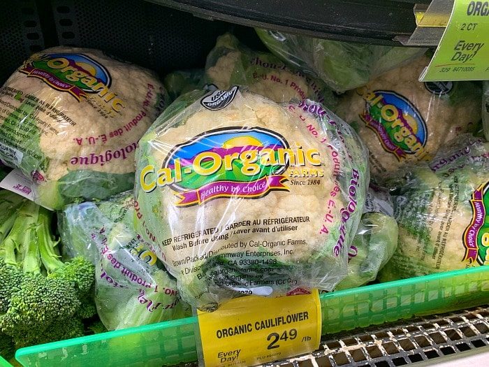 Packages of Cal-Organic Farms organic cauliflower are displayed in a grocery store, solid for your next air fryer cauliflower recipe. Each one is wrapped in branded plastic packaging, with a price tag showing $2.49 per pound. Broccoli is partially visible in the display.