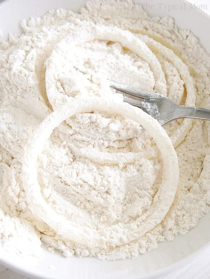 Crispy fried onions being coated in flour with a fork in a white bowl.