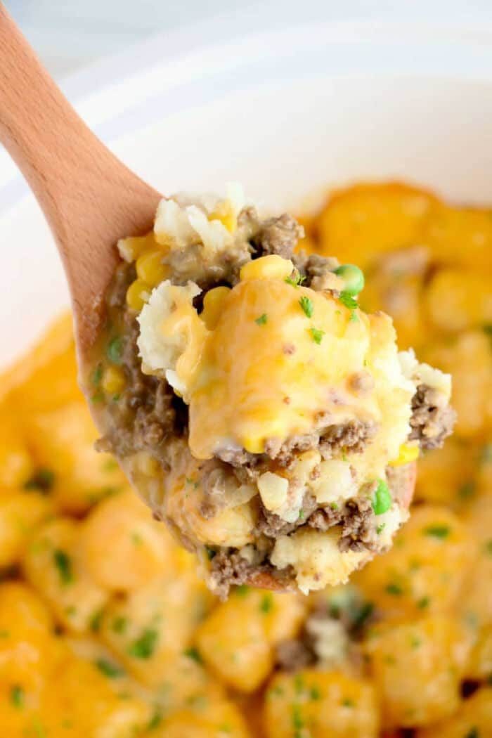 A wooden spoon holds a serving of crock pot tater tot casserole with cheesy ground beef, peas, and corn.