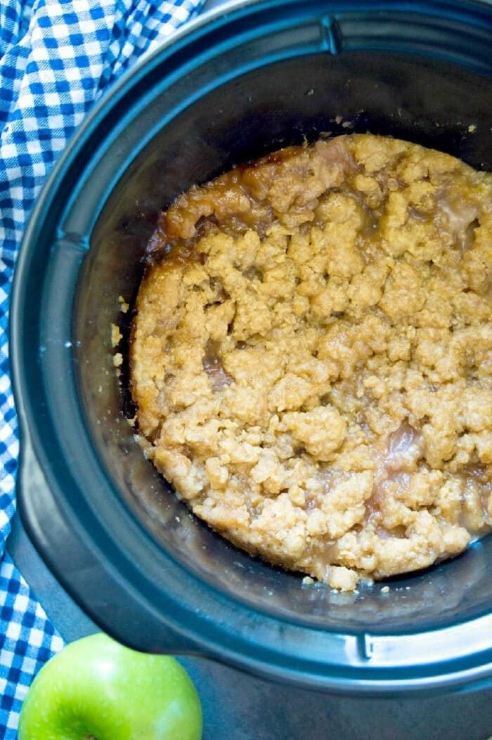 A crockpot filled with warm crockpot apple crisp sits beside a green apple and a blue checkered cloth.