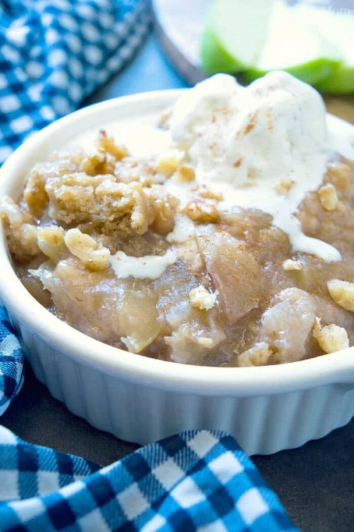 Crockpot apple crisp topped with a scoop of vanilla ice cream in a white dish, next to a blue checkered cloth.