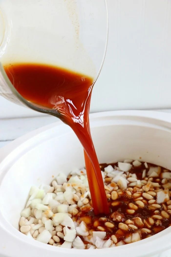 Pouring sauce into a slow cooker with chopped onions and white beans creates the solid base for a homemade crockpot baked beans recipe.