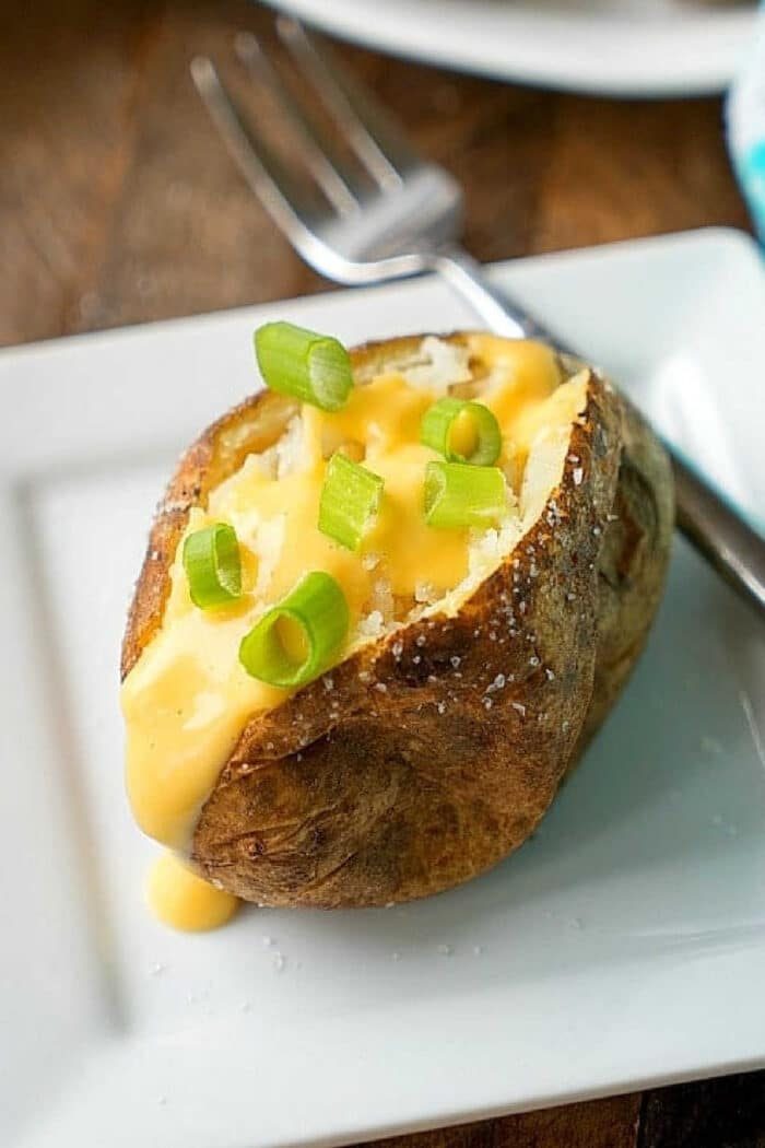 Crockpot baked potatoes topped with cheese sauce and chopped green onions on a white plate with a fork.