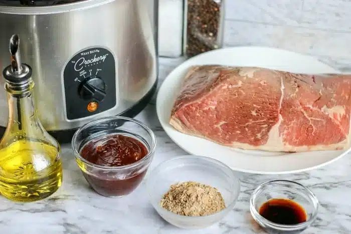A raw beef roast, oil, seasonings, sauce, and a slow cooker on a marble countertop—everything you need to make savory crockpot bbq beef.