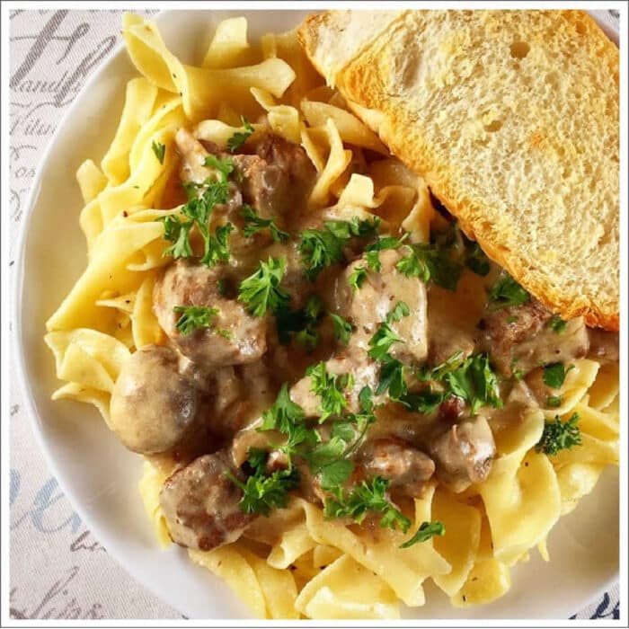 A plate of creamy crockpot beef stroganoff over egg noodles, topped with parsley and served with a slice of bread.