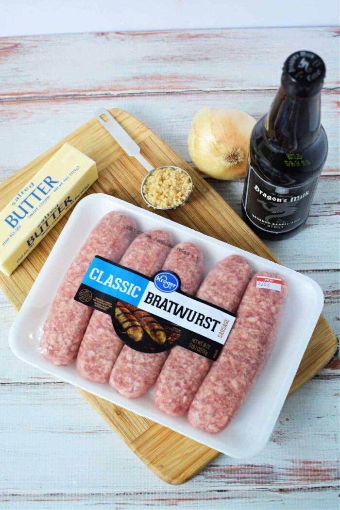 bratwurst, butter, chopped garlic, onion, and a beer bottle rest on a cutting board—solid for crafting good Crockpot Brats.