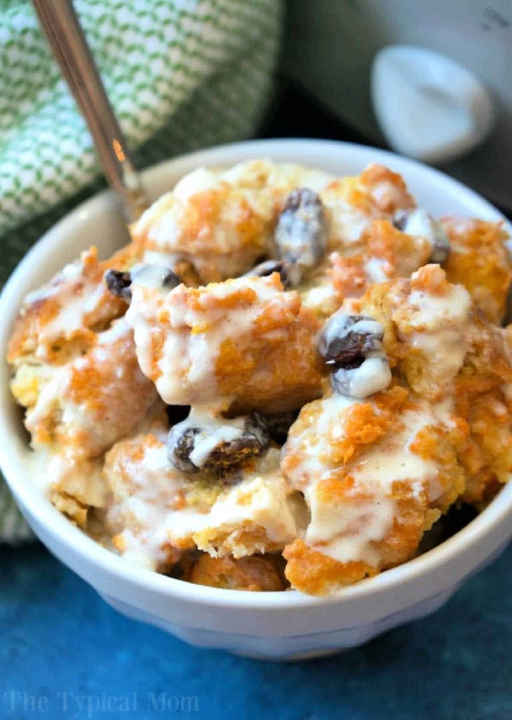 A bowl of sweet crockpot bread pudding with raisins, topped with creamy icing or sauce, sits beside a green patterned cloth.