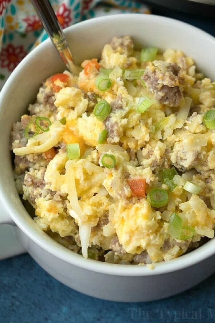 Bowl of scrambled eggs with sausage, cheese, potatoes, and chopped green onions, with a spoon—like the comforting flavors you'd find in a classic crockpot breakfast casserole recipe.