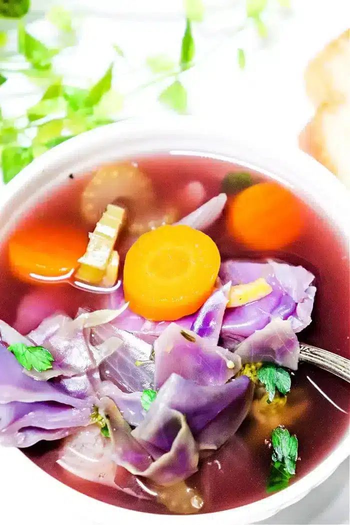 A bowl of Crockpot Cabbage Soup contains purple cabbage, sliced carrots, celery, and herbs in a light broth. A spoon is partially submerged in the soup. Bright green leafy garnishes are visible in the background.