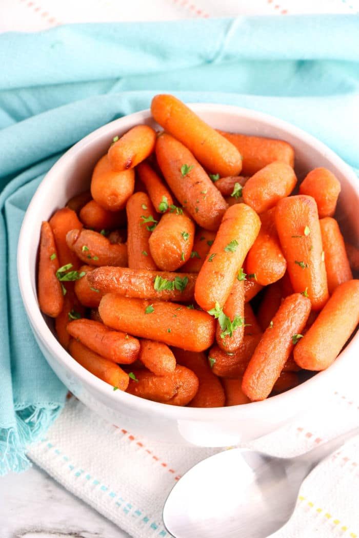 A bowl of tender crockpot carrots garnished with parsley sits on a blue cloth, with a spoon nearby.