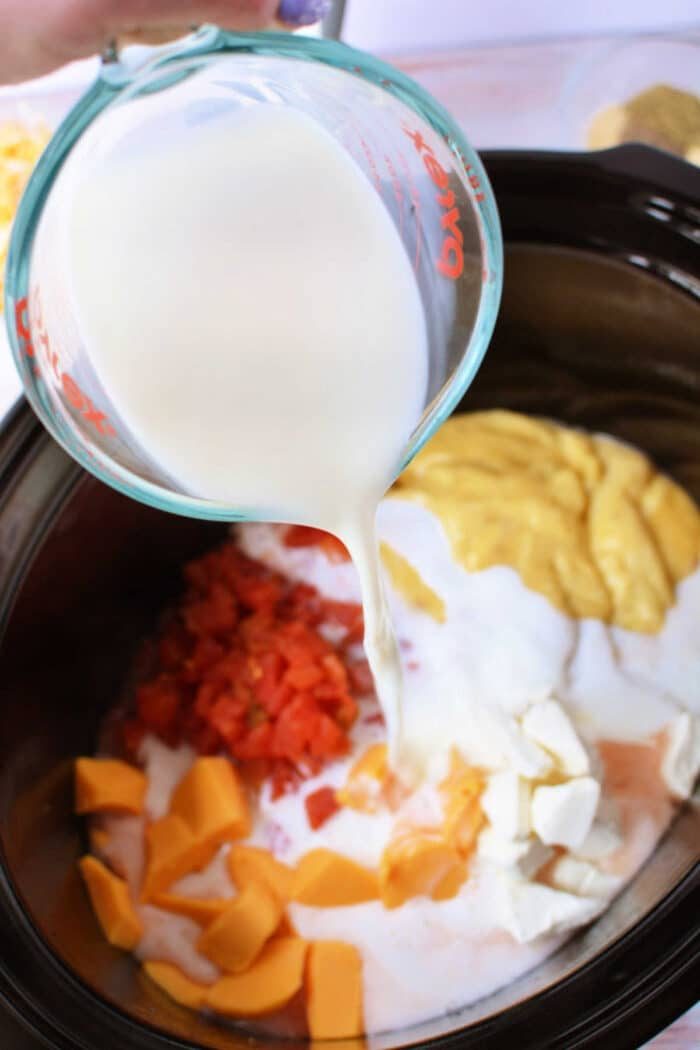 A measuring cup pours milk into a slow cooker filled with cheese, diced tomatoes, and all the good ingredients needed for Crockpot Chicken Spaghetti.